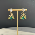 Gold earring stand with green and gold bee-shaped earrings on a marble surface.